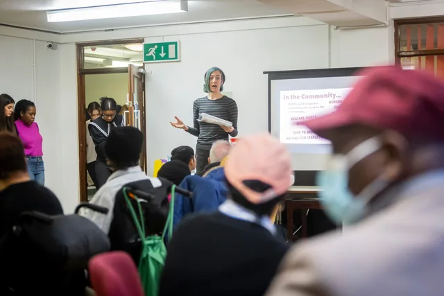 A diverse group attending a community meeting with a presenter speaking in front of a screen. Community roles and operating routines
